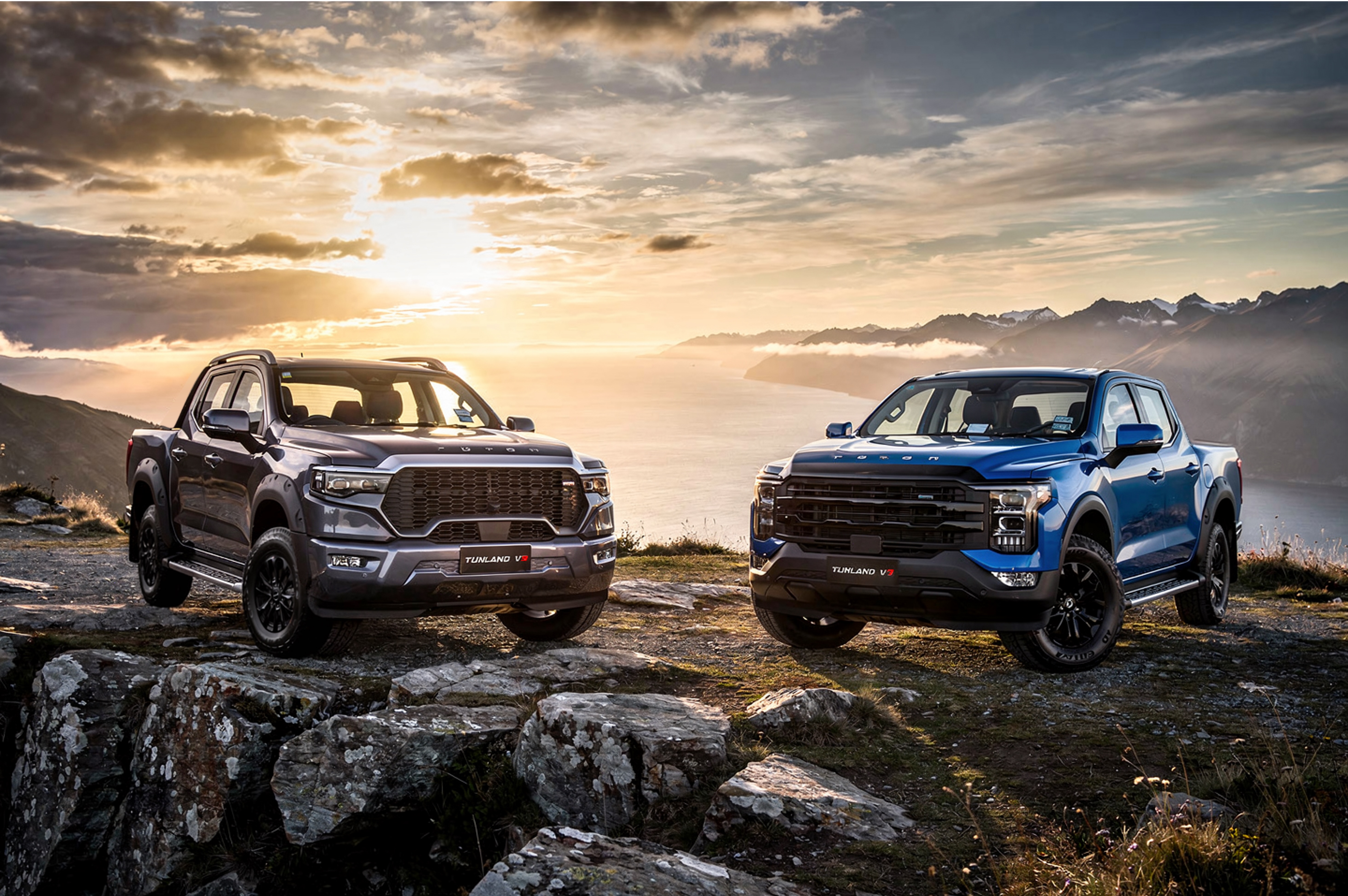 Two Foton Tunland V9 utes, one grey and one blue, parked on a rocky coastal ridge at sunrise overlooking the sea and mountains